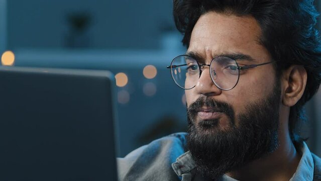 Thoughtful arabian indian bearded adult man thinking of problem solution working on laptop at home doubtful male face in glasses looking at computer screen watching video late night darkness overtime