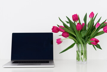 Laptop with empty black screen and bouquet of pink tulip flowers in a transparent vase on a white table. Working space. Home office
