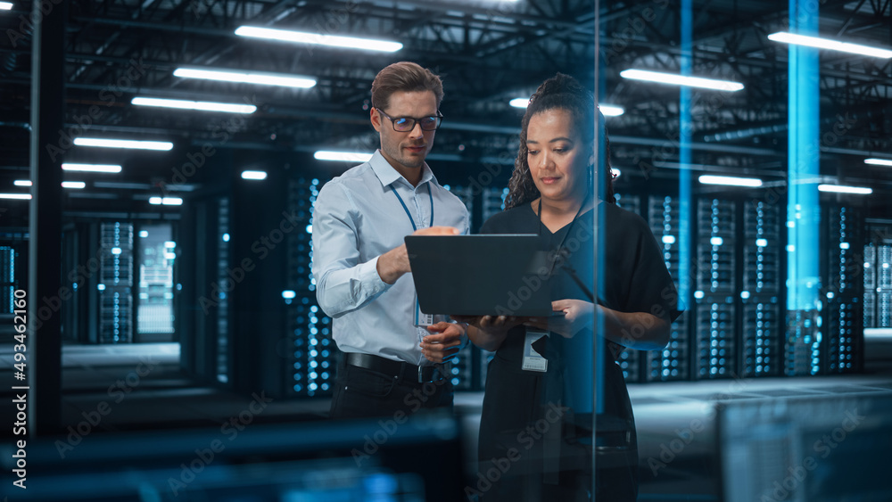 Poster Multiethnic Female System Administrator and Caucasian Male Information Technology Engineer Discussing and Brainstorming with Laptop while Work in Tech Facility Data Center