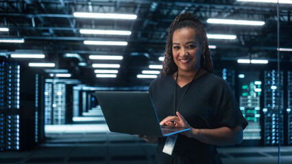 African American Female IT Specialist Using Laptop Computer, Standing in Big Warehouse Data Center....