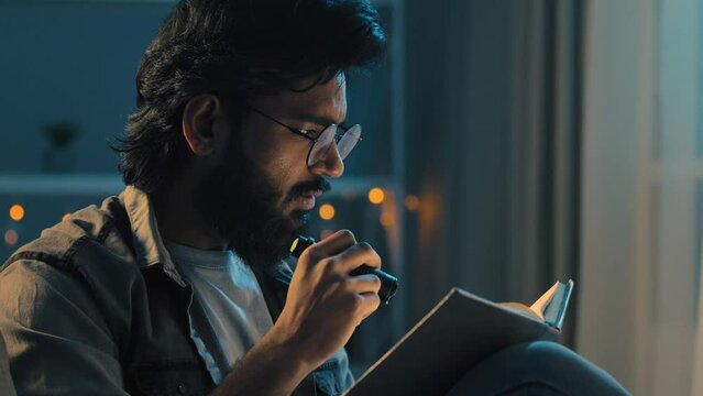 Arab indian adult man with beard wearing glasses sitting at home night late time darkness blackout reading spiritual book textbook with flashlight home schooling studying learning preparing for exams