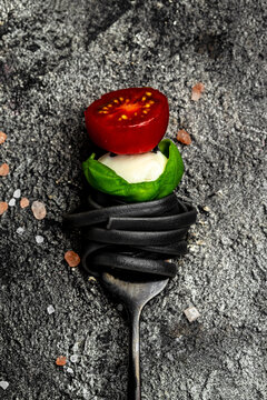 black pasta caprese salad tomatoes, mozzarella and green basil leaves on a black fork, Pasta with cuttlefish ink. vertical image. top view. place for text