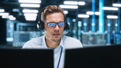 Portrait of a Young Caucasian Male Call Centre Worker Wearing a Phone Headset Talking at the Support Line with Serious Face in a Modern Open Plan Office at Night. Helpline Concept - obrazy, fototapety, plakaty
