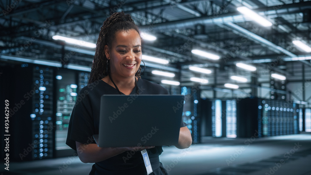 Wall mural Multiracial Data Center IT Technician Standing at the Server Rack Corridor with a Laptop Computer. She is Visually Inspecting Something while Looking Away. Night Office Concept