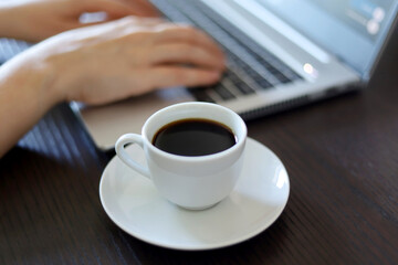 Coffee cup with saucer on background of female hands on laptop keyboard. Woman works sitting at...