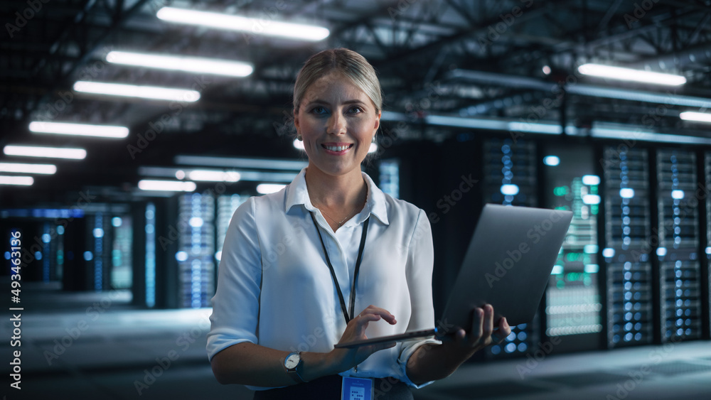 Poster cheerful blonde female it specialist using tablet computer in data center, while smiling to the came