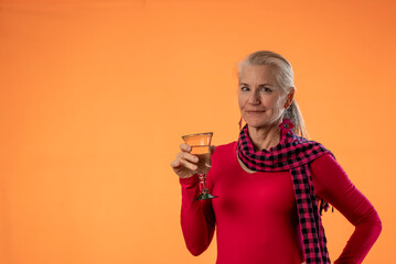Beautiful smiling happy blond mature woman holding wineglass and drinking wine on solid orange background with copy space sitting on stool.