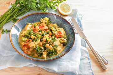 Curry dish from chickpeas, broccoli, bell pepper and chicken meat on a blue plate, served with cutlery, herbs and napkin on a light wooden table, copy space, selected focus