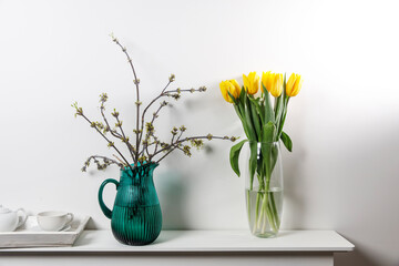 Bouquet of yellow tulip in the glass vase and American maple branches in green glass jar on white table