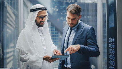 European Businessman Talking with Arab Investment Partner while Riding Glass Elevator to Office in a Modern Business Center. International Corporate Associates Use Tablet Computer in a Lift. © Gorodenkoff