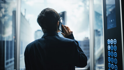 Successful Stylish Japanese Businessman Riding Glass Elevator to Office in Modern Business Center. Handsome Happy Man Talking on a Phone, Taking an Important Business Call in a Lift. © Gorodenkoff