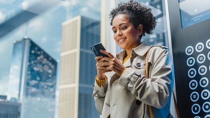 Successful Latin American Female Riding Glass Elevator to Office in Modern Business Center. Beautiful Manager Smile while Using Smartphone, Write Text Message, Check Social Media and Emails in a Lift.