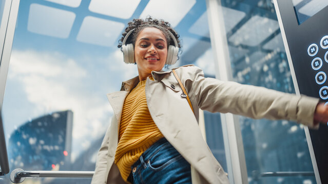 Carefree African American Female Riding Glass Elevator To Office In Modern Business Center. Beautiful Multiethnic Latin Woman Is Happy, Smile, Listen To Music On Headphones And Dance In The Lift.