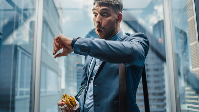 Business Manager is Late for Work, Riding Glass Elevator in Modern Office Building. Handsome Man Eating a Tasty Take Away Hamburger in a Hurry on the Way to Corporate Meeting in a Lift.