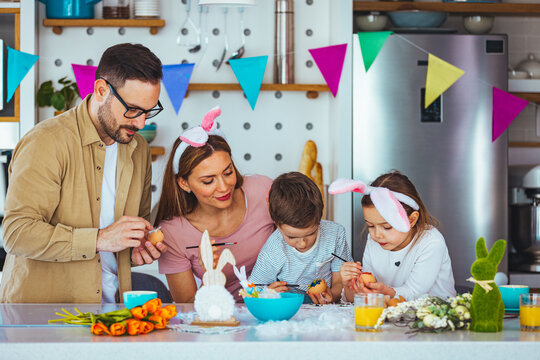 Joyful family wearing bunny ears headbands gathering at table in modern light kitchen and paining Easter eggs together. Happy family spending Easter together in the kitchen