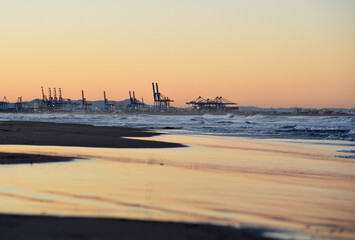 Fototapeta premium Coastline near sea port with quay crane on sunset. Gantry cranes in seaport on sunrise. Waves in sea at coast on sunset. Shore of beach overlooking ship port. Seascape in Spain. No people on beach.