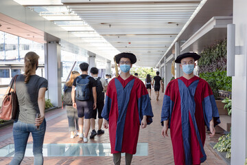 two happy proud Asian PhD graduated male students in Academic dress gown and face masks walk in campus