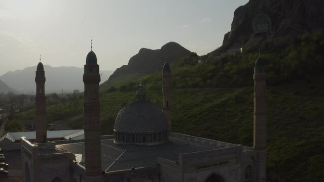 A beautiful Muslim mosque in rays of sunset sunlight against background of rocky mountains and sky. Flying a quadrocopter around a mosque in an urban environment in golden sunlight. Kyrgyzstan osh