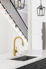 A kitchen sink detail shot with a gold faucet, grey island, white marble countertop, and lights hanging above the island.