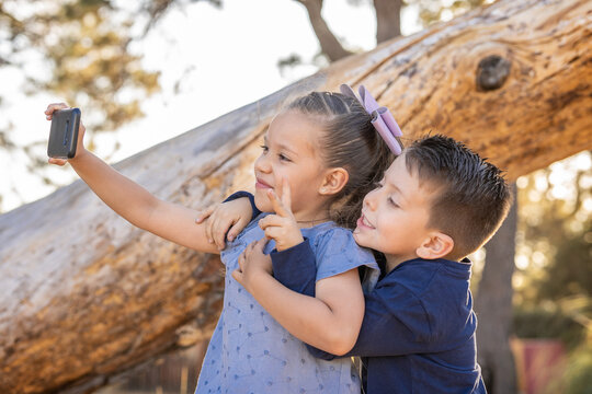 Niños felices hermanos amigos amistad tomándose fotos fotografías selfies retrato con el celular alegres jugando y disfrutando en el parque al aire libre atrás de un tronco de un árbol