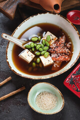 Bowl of miso soup with salmon fillet, edamame beans and tofu cheese, vertical shot on a brown stone background