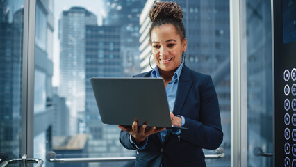 Fototapeta premium Successful Black Female Riding Glass Elevator to Office in Modern Business Center. Beautiful African American Manager Using Laptop Computer to Check Schedule, Social Media and Work Emails in a Lift.