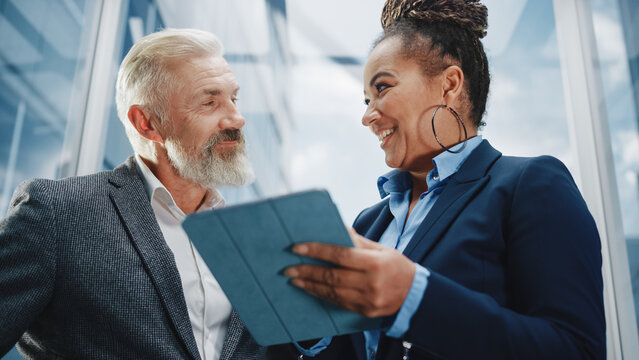 Middle Aged Businessman Talking with International Investment Partner while Riding Glass Elevator to Office in a Modern Business Center. Corporate Associates Use Tablet Computer in a Lift.