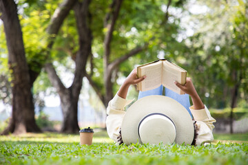 Woman reading book in grass under tree with cup of coffee. Relax in summer time holiday laying on the grass field comfortable feeling.