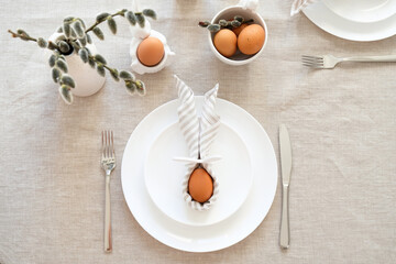 Easter dinner table setting with egg in Easter bunny napkin on plate, fork and knife, willow on linen tablecloth. Flat lay, top view.
