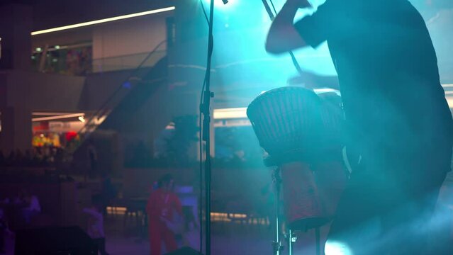 Man with ponytail dreadlocks standing back to camera playing ethnic drums on stage in smoke and spotlights. performance takes place in the lobby of the shopping mall. Rear view medium shoot 