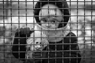Little refugee girl with a toy behind a metal fence. Social problem of refugees and internally...