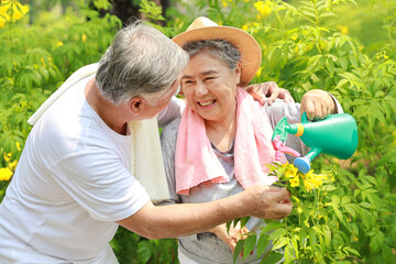 Asian senior couple incasual embracing and spending time in garden backyard while watering flowers plants with laughing and smiling on sunny day after retired. Happy elderly outdoor lifestyle concept.