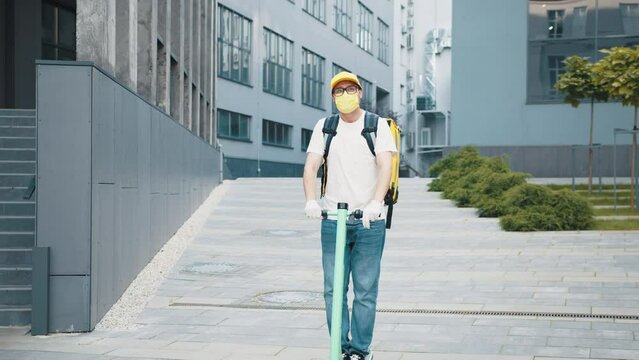 Delivery man with yellow backpack and protective mask riding electric scooter through the city with food delivery, business center background. Excellent delivery during quarantine. Concept Delivery