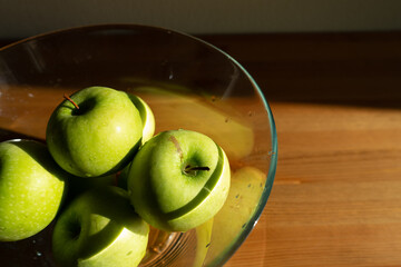 green apples in a transparent plate on a wooden table in the interior. kitchen table in sunlight.