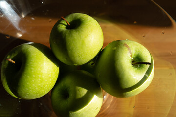 green apples in a transparent plate on a wooden table in the interior. kitchen table in sunlight.