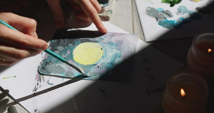 a girl paints a painting in the sun, gray strokes of paint on a spotted picture, a brush runs oil acrylic paints on a small canvas, impressionism burns in the shadow of a candle, a drawing of the moon