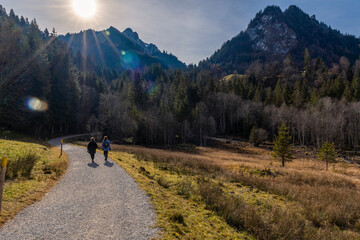 Two women walk on a winding dirt road between fields and mountains in late autumn on a sunny day in the Schwarzee region of Canton Fribourg, Switzerland.