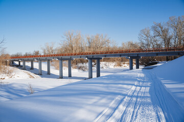 A bridge over a frozen river against a background of white snow. A road across a bridge in a remote forest. Snowmobile footprints in the snow. Winter landscape.