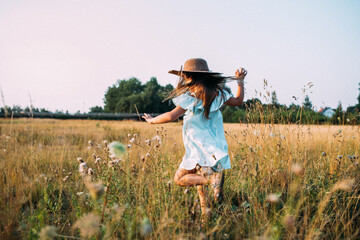 Kind mit langen Haaren in hellblauen Kleid und Hut rennt freudig über eine Blumenwiese im Sommer