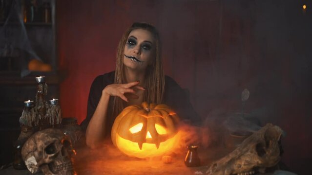 Come here. Halloween concept. Black witch holding Halloween pumpkin with carved smily face sitting on the table dark room inviting with hand gesture. Jack o lantern head. Looking at camera