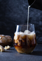 Cream from a black milk jug is poured into iced coffee. Cookies and coffee on a dark background