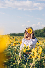 Obraz na płótnie Canvas Beautiful young long-haired blonde woman in a white blouse with bare shoulders holds a sunflower in her hands on a sunflower field.Summer concept.