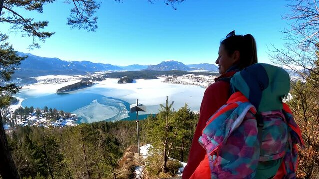 Active woman with a backpack walking to the edge of the viewing platform of Tabor, Taborhoehe with scenic view on the frozen Faak lake in the Karawanks in Carinthia in Austrian Alps. High mountains
