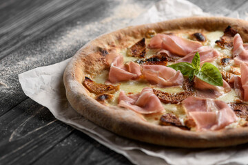 Delicious freshly baked meat pizza. Appetizing whole-wheat pizza with prosciutto and figs. Served on parchment paper. Dark wooden background with sprinkled flour. Cropped image, selective focus.