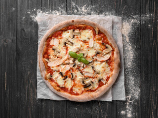 Top view of delicious freshly baked pizza. Appetizing pizza from whole grain dough with cheese, mushrooms and chicken meat. Served on parchment paper. Dark wooden background with sprinkled flour.
