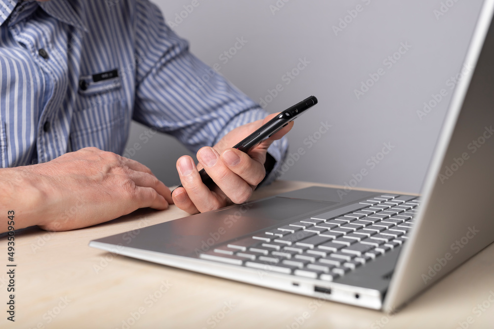 Poster Man sitting at table with laptop and holding phone. Checking information, reading or typing message while working on computer. Man hands closeup. High quality photo