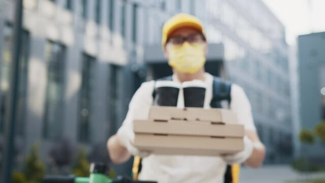 Handsome delivery man in protective face mask holding stack of pizza cardboard boxes with two cups of coffee. Unrecognizable Male courier with pizza looking at camera