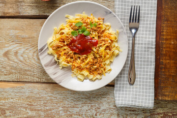 Pasta bolognese garnished with herbs and cheese in a plate on a wooden table next to a fork and a napkin.