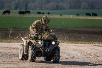British army soldier driving a small ATV quad bike across open countryside on a military exercise