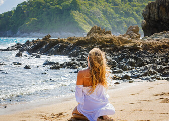 A young girl in a white dress sits on the sand by the sea and looks into the distance. High quality photo